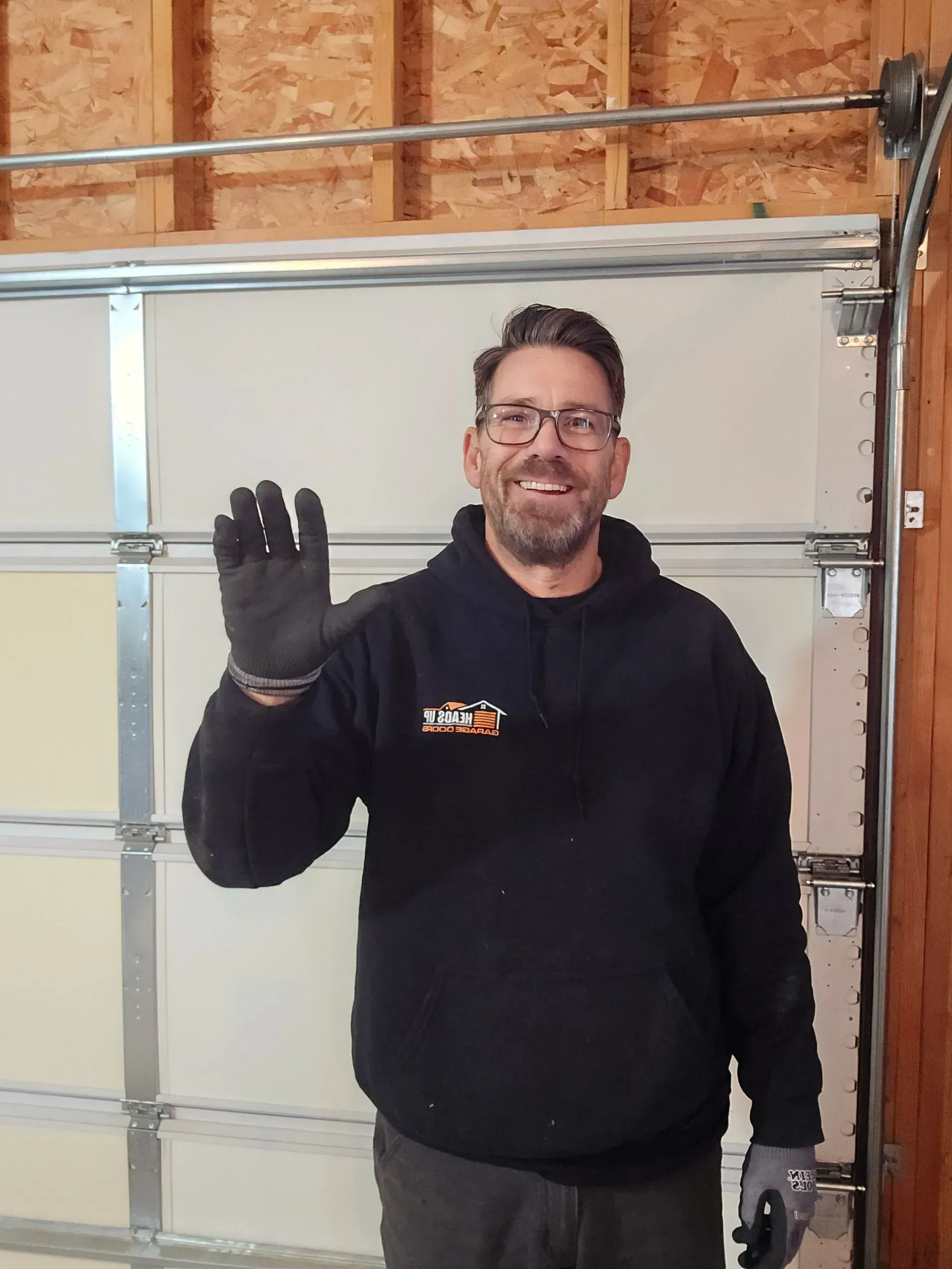 Man in a black hoodie waves in front of a garage door. He wears glasses and work gloves.