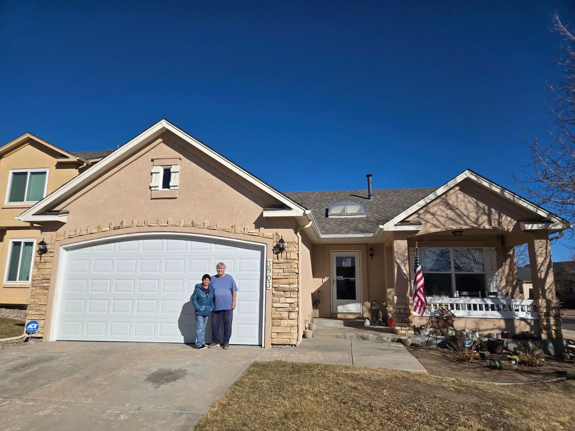 Two people standing in front of a tan house with a white garage door, bright blue sky overhead.