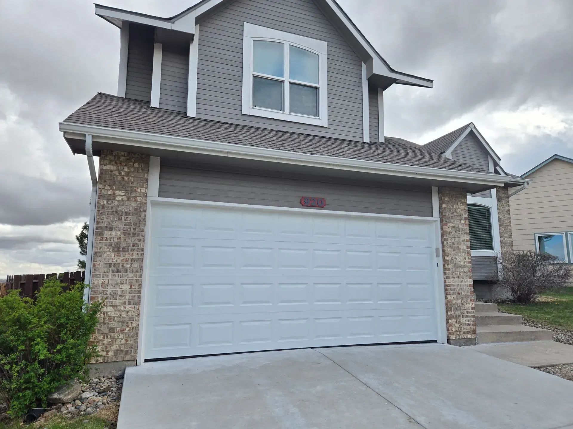 Two-story house with white garage door, tan stone accents, gray siding, and a concrete driveway under a cloudy sky.