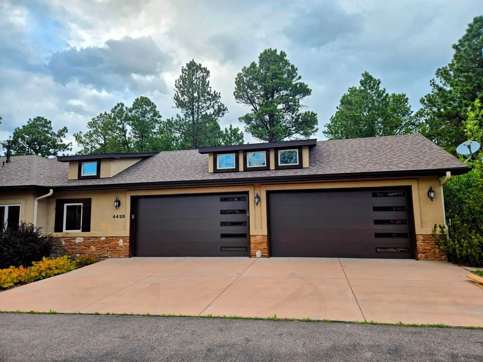 Two-car garage with brown doors, beige siding, and a concrete driveway. Trees and cloudy sky in background.