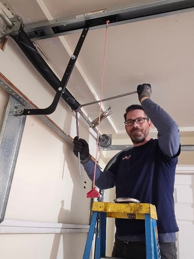 Man on a ladder repairing a garage door opener. He wears gloves and smiles. Indoors, against a white wall.