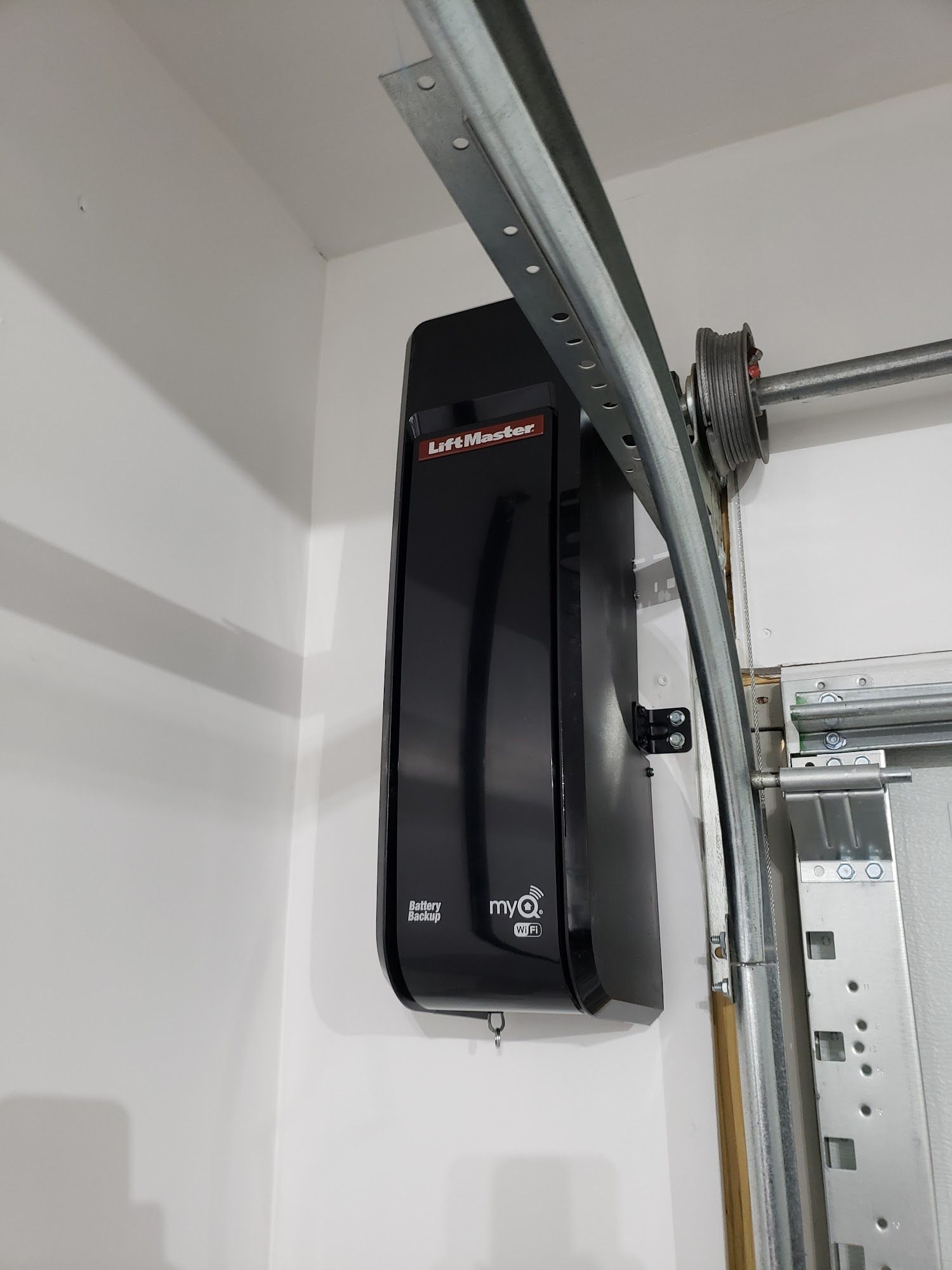 Black garage door opener mounted on a white wall; metal track and wheel visible.