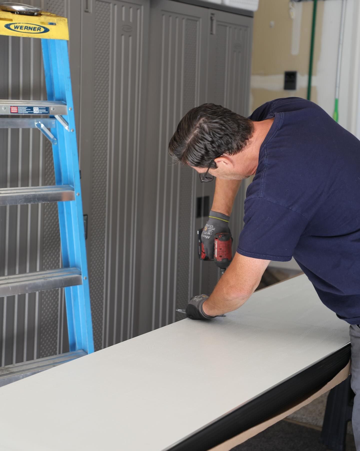Man using a power tool to cut a white board in a garage; a ladder is to the left.