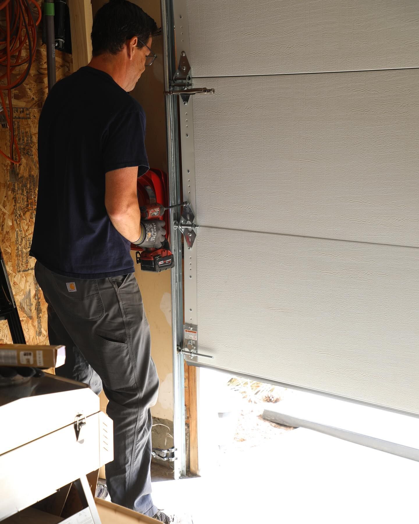 Man using power drill on a garage door, standing on a workbench in a garage.