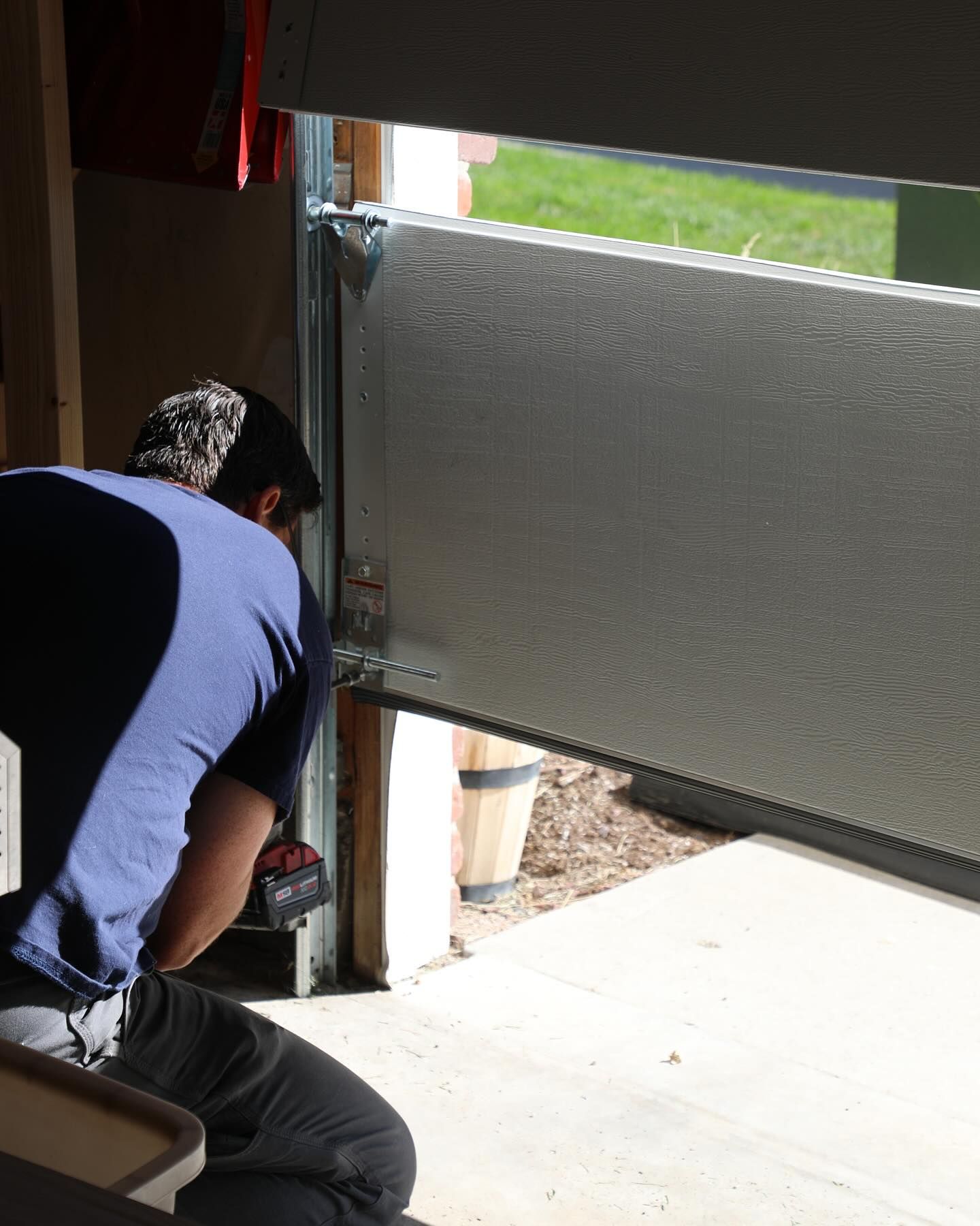 Person installing a gray garage door. He is using a drill while kneeling. Sunlight shines in.