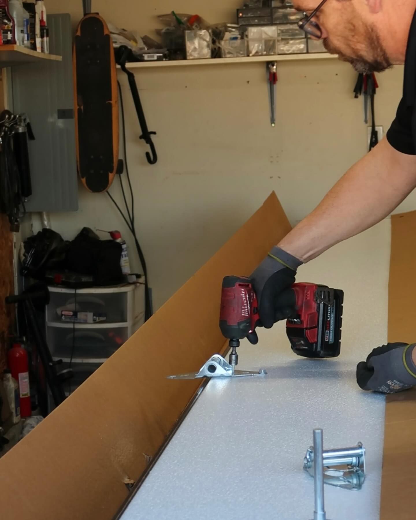 Person using a red cordless shear to cut a metal sheet in a garage, holding the tool with a gloved hand.