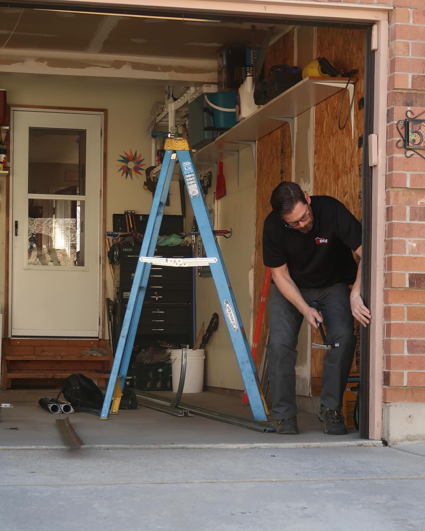 Man working on a garage door frame, using a tool. He stands near a ladder, with tools and shelves visible.