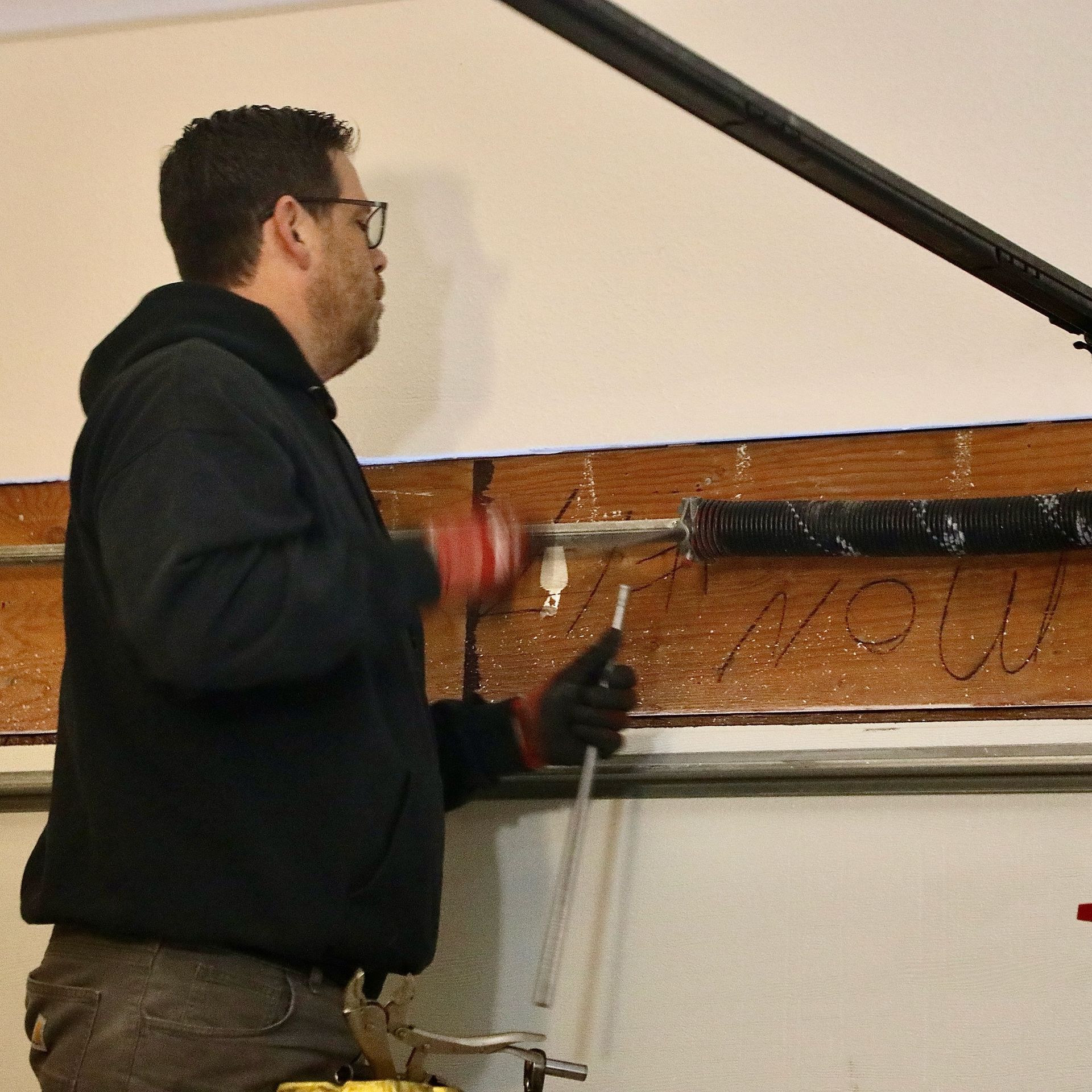 Man repairing garage door spring, wearing black hoodie, gloves, and glasses.