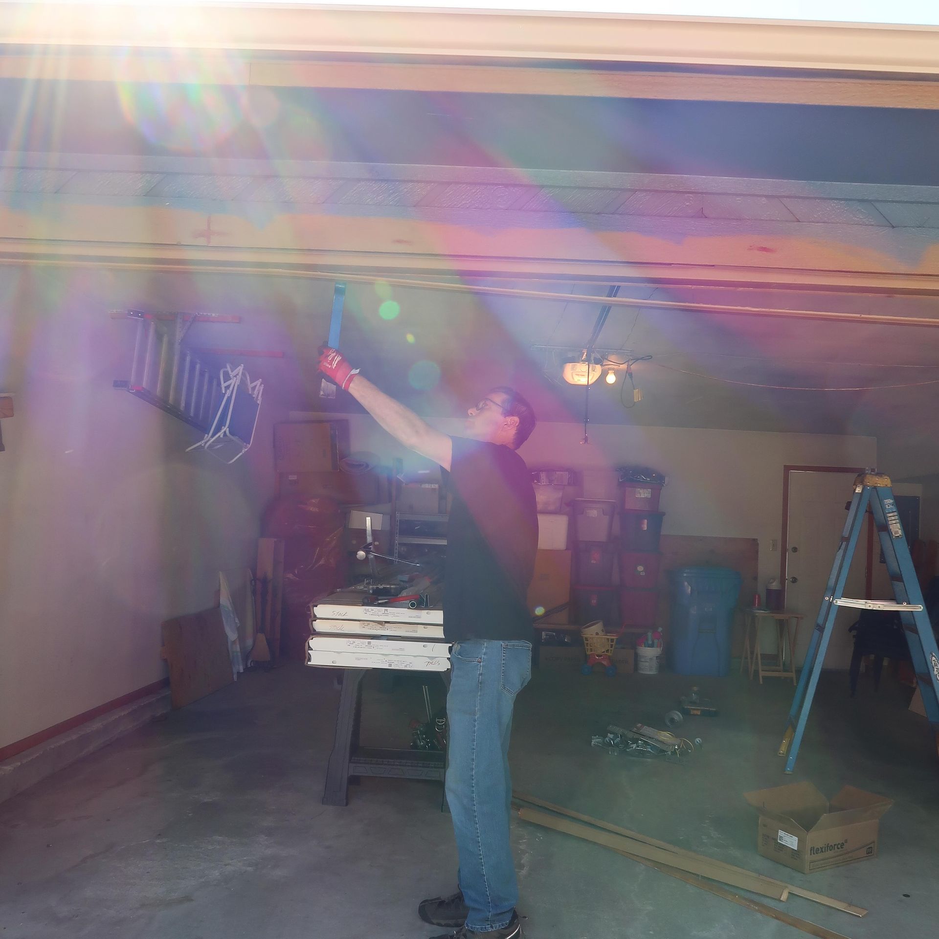 Person in a garage, raising arm with tool towards ceiling. Bright sunlight.