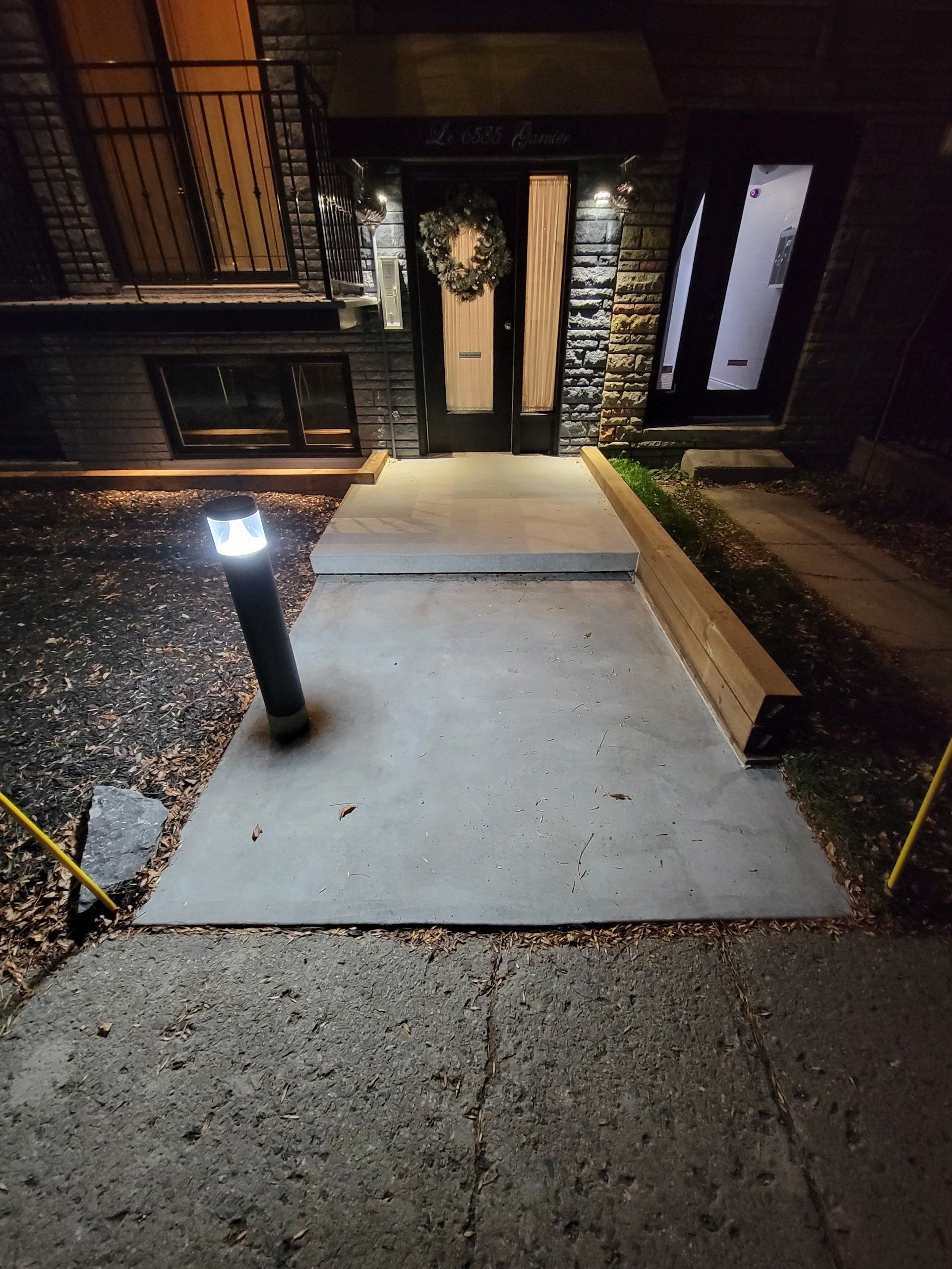 Allée en béton menant à une maison dont la porte d'entrée est éclairée. Extérieur sombre avec éclairage de cheminement.