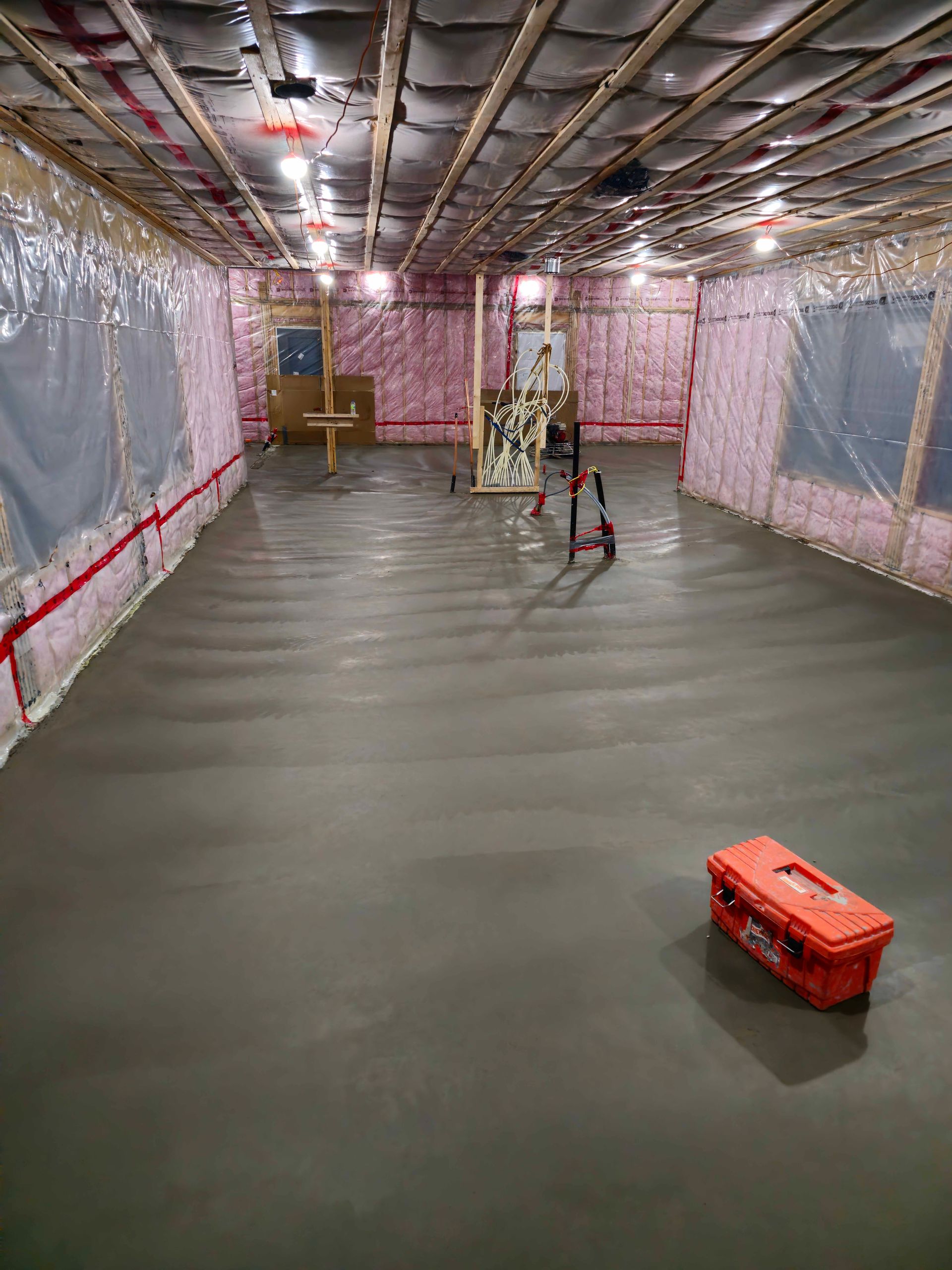 Plancher de sous-sol en béton fraîchement coulé, avec isolation et montants apparents. Boîte à outils rouge dans un coin.