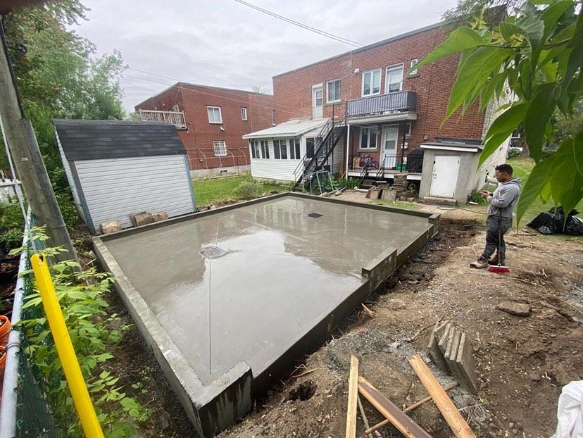 A man is standing in front of a concrete foundation in a backyard.