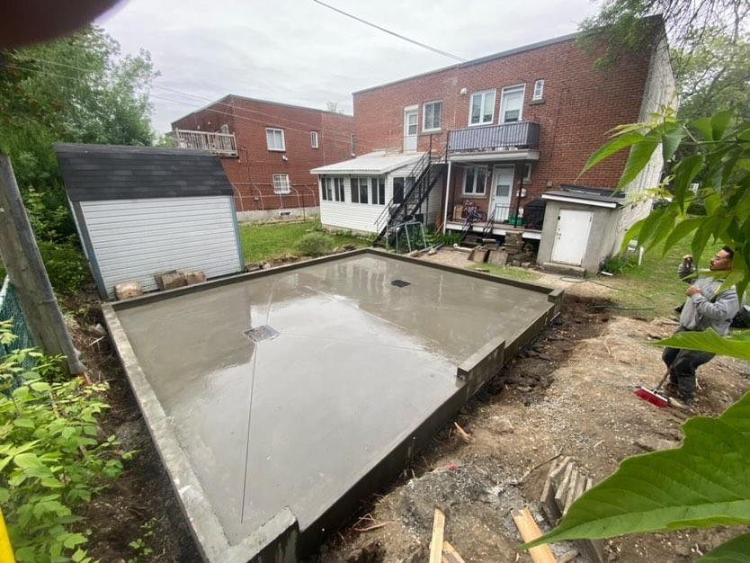 Base en béton fraîchement coulée dans une cour arrière ; bâtiments en briques à deux étages en arrière-plan.
