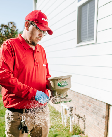 Person in red uniform using a fertilizer spreader beside a house.