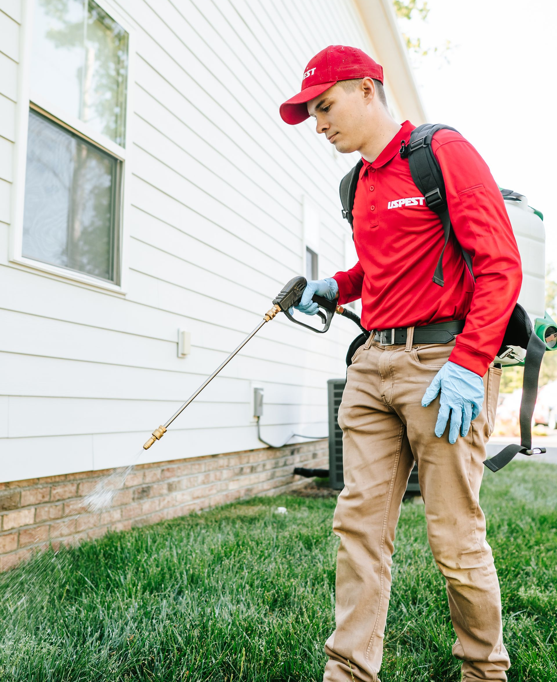 Worker in red uniform spraying pesticide along a house foundation with a backpack sprayer