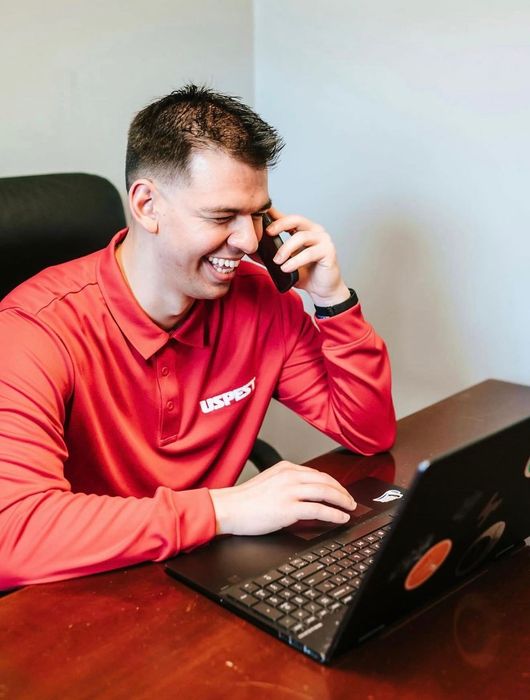 Man in a red shirt smiling while talking on a phone beside a laptop at a desk