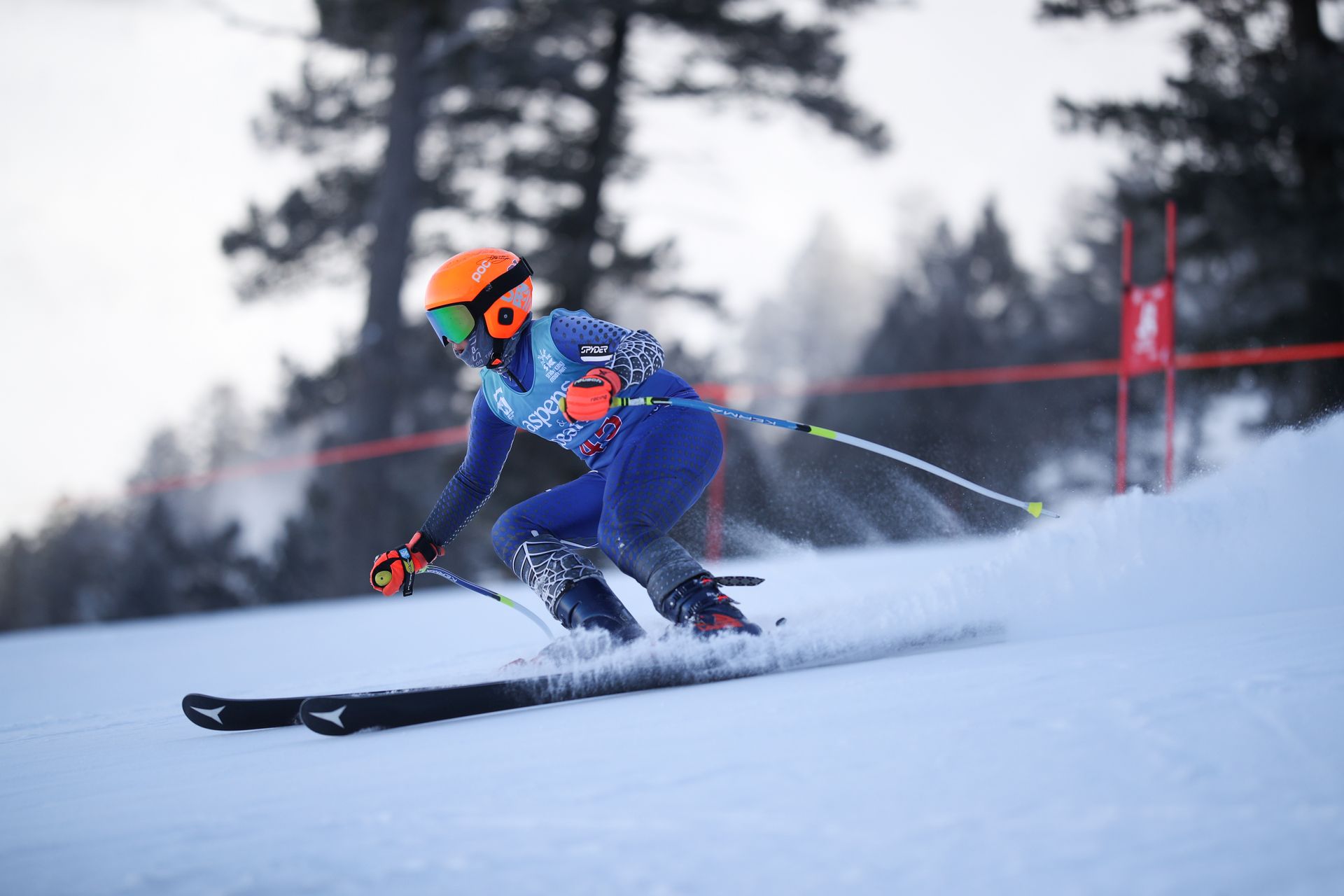 Skier in blue and orange racing downhill on snow, leaning into a turn with ski poles raised