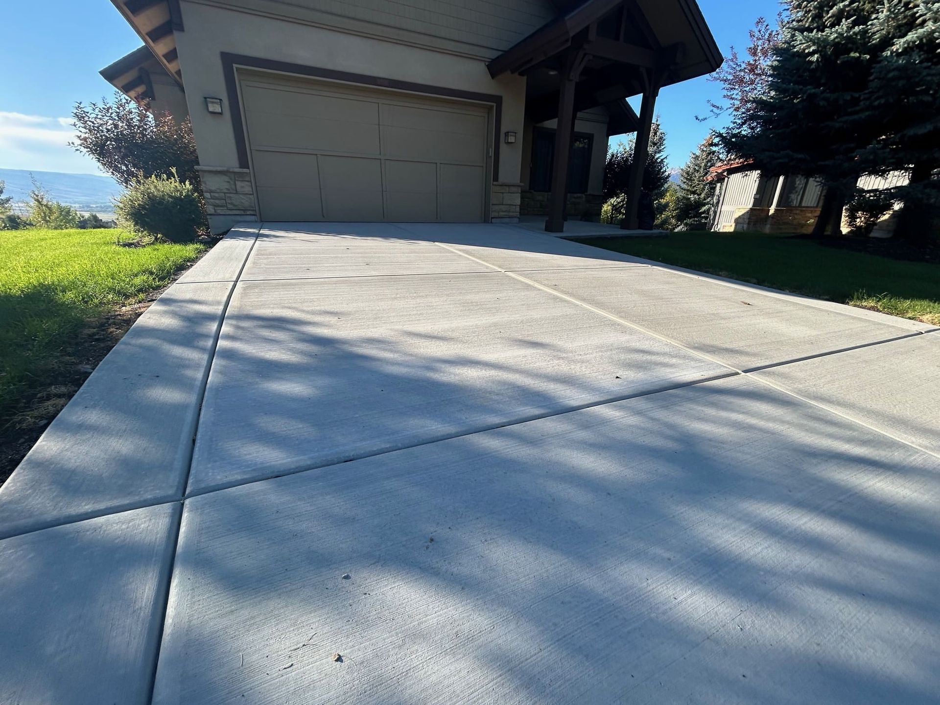 Concrete driveway leading to a house with a garage, green lawn, and trees. Sunny day.