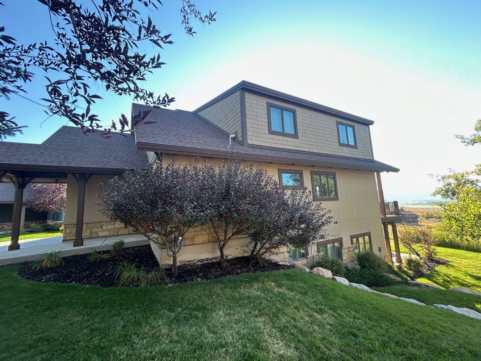 Two-story beige house on a grassy hill, with a covered walkway and lush greenery.
