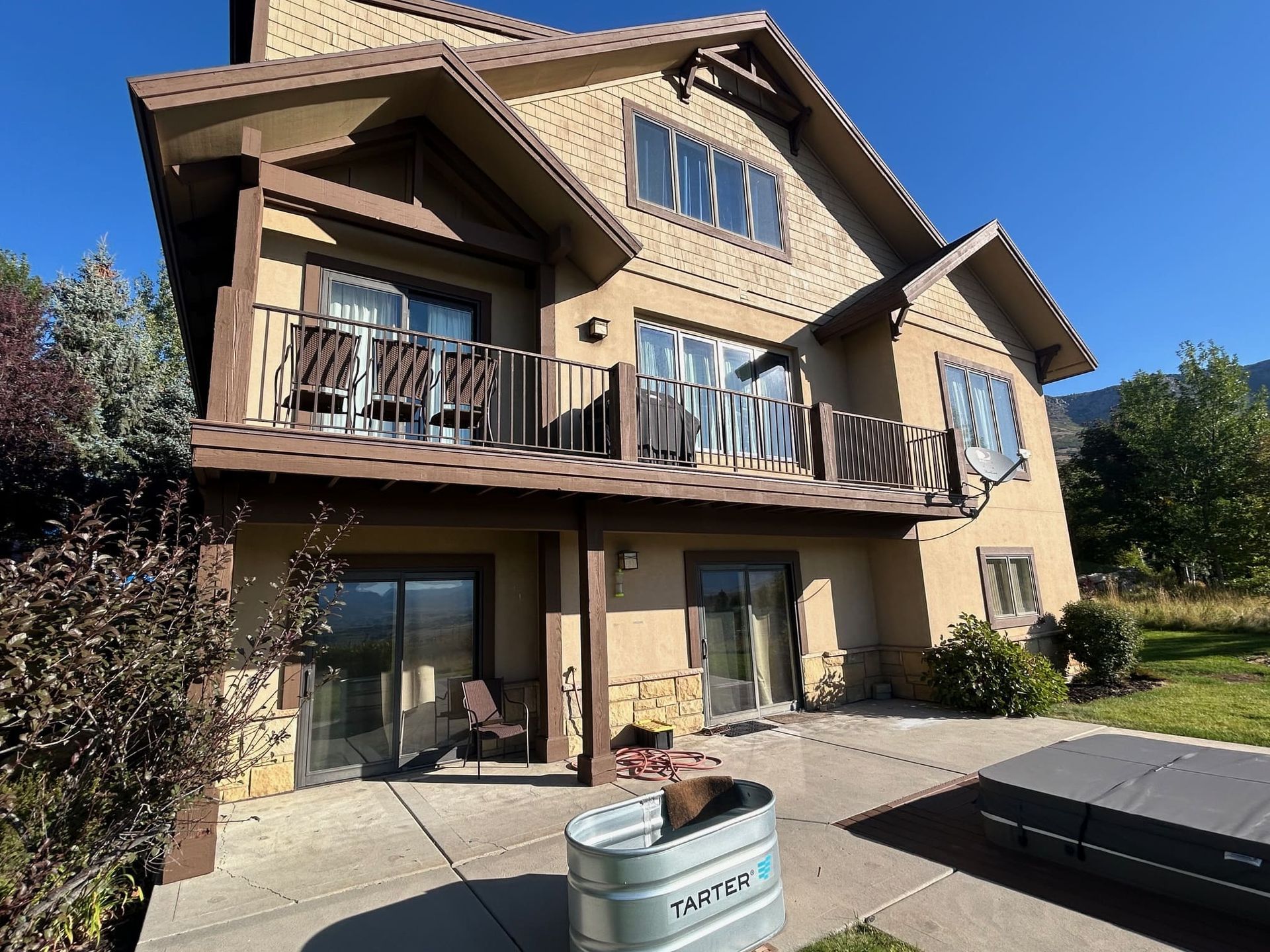 Two-story tan house with balconies, stone accents, and a patio, set against a mountain backdrop.