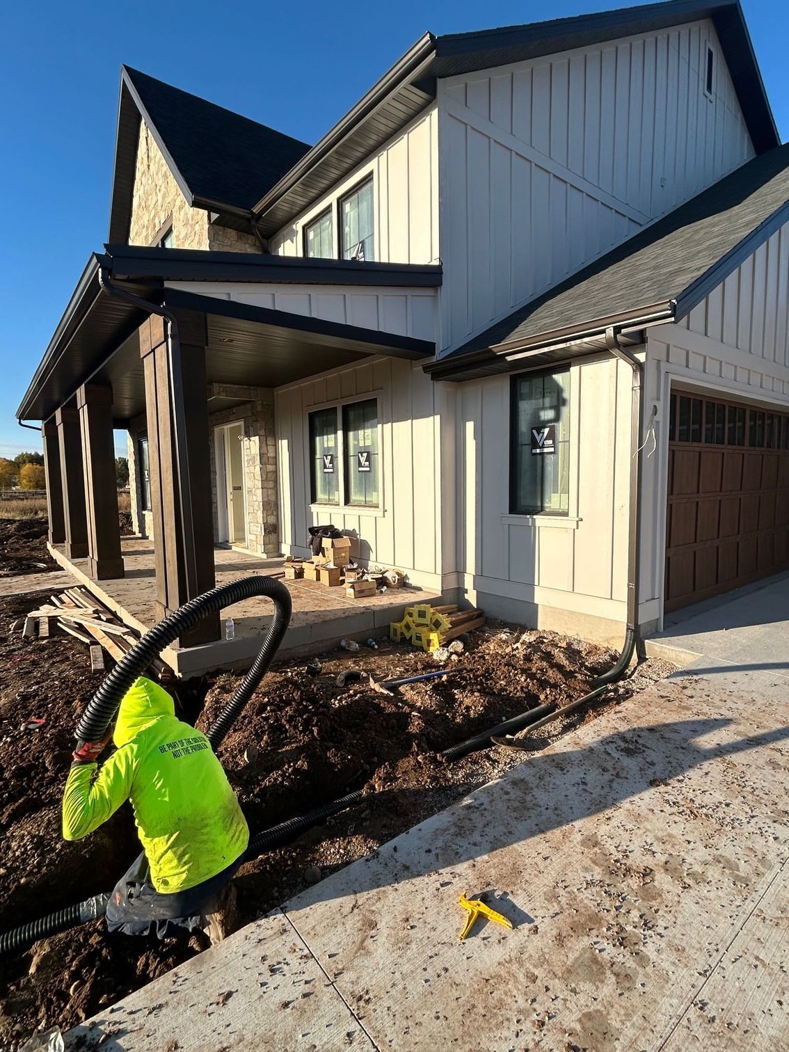 Person in neon jacket working on a black pipe near a new house with white siding and stone accents.