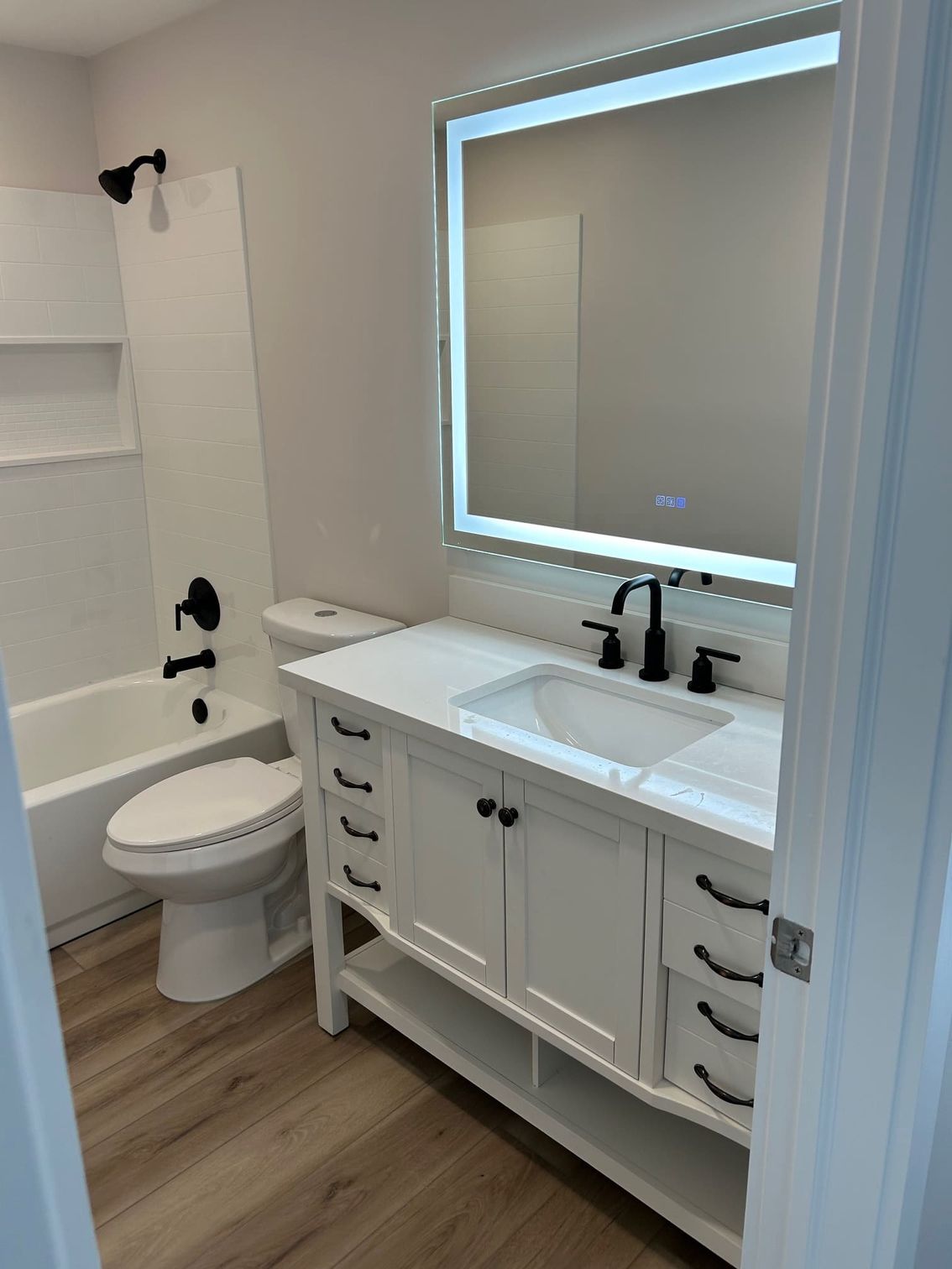 White bathroom with vanity, toilet, and bathtub. Black fixtures, large mirror, and wood-look flooring.