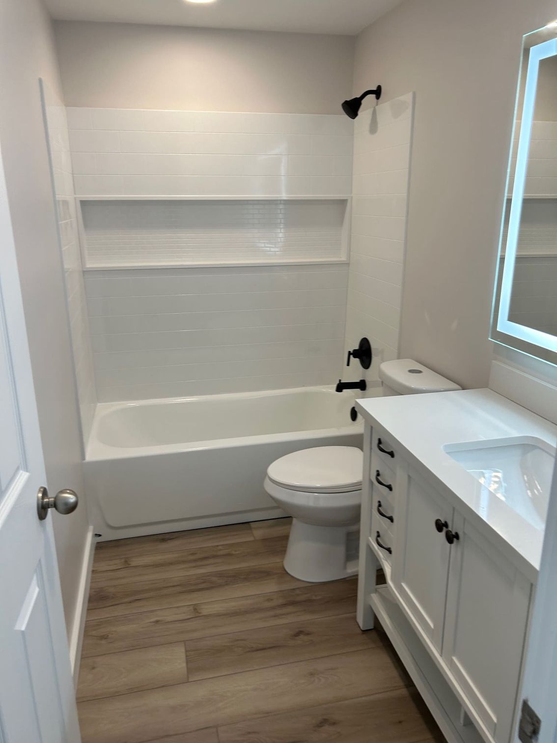 Bathroom with white tub, vanity, and toilet; black fixtures; light wood-look floor.