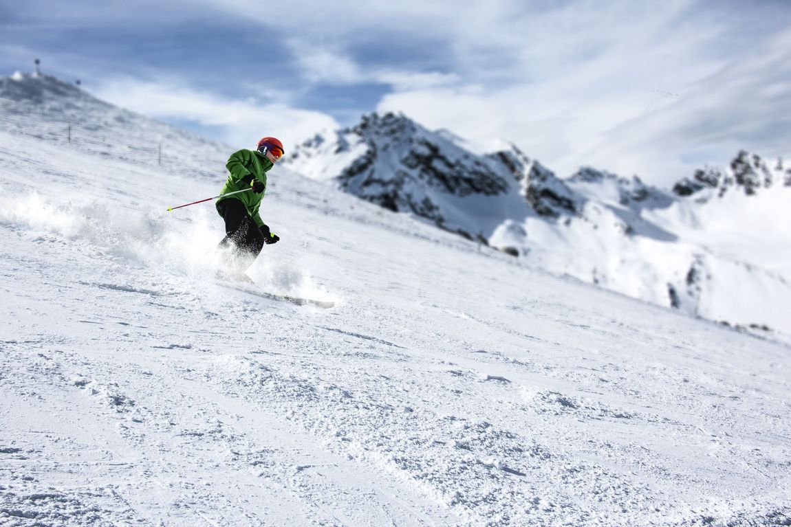 Skier in green jacket carves down snowy mountain slope with jagged peaks in background.
