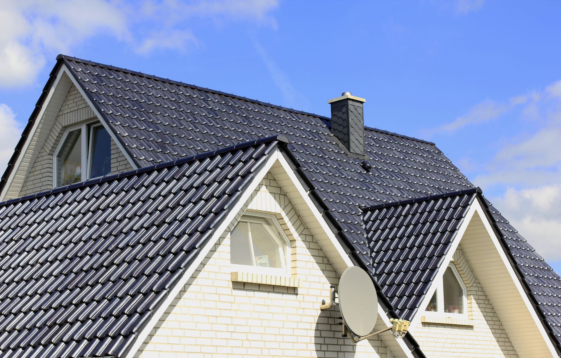 White brick house with dark gray tiled roof, dormers, chimney, and satellite dish against a blue sky.