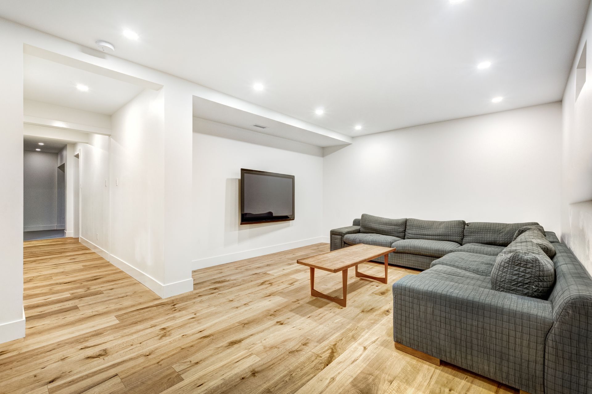 Modern living room with wooden floor, gray sectional sofa, TV, and coffee table. White walls and recessed lighting.