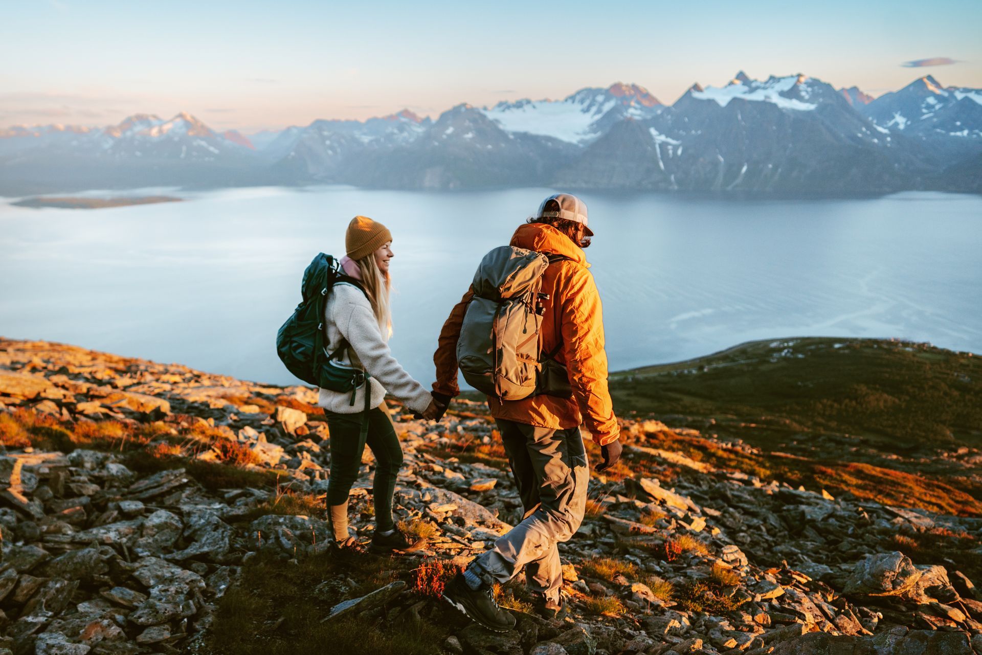 Couple hiking hand-in-hand on a mountain, overlooking a lake and mountains. Golden hour sunlight.