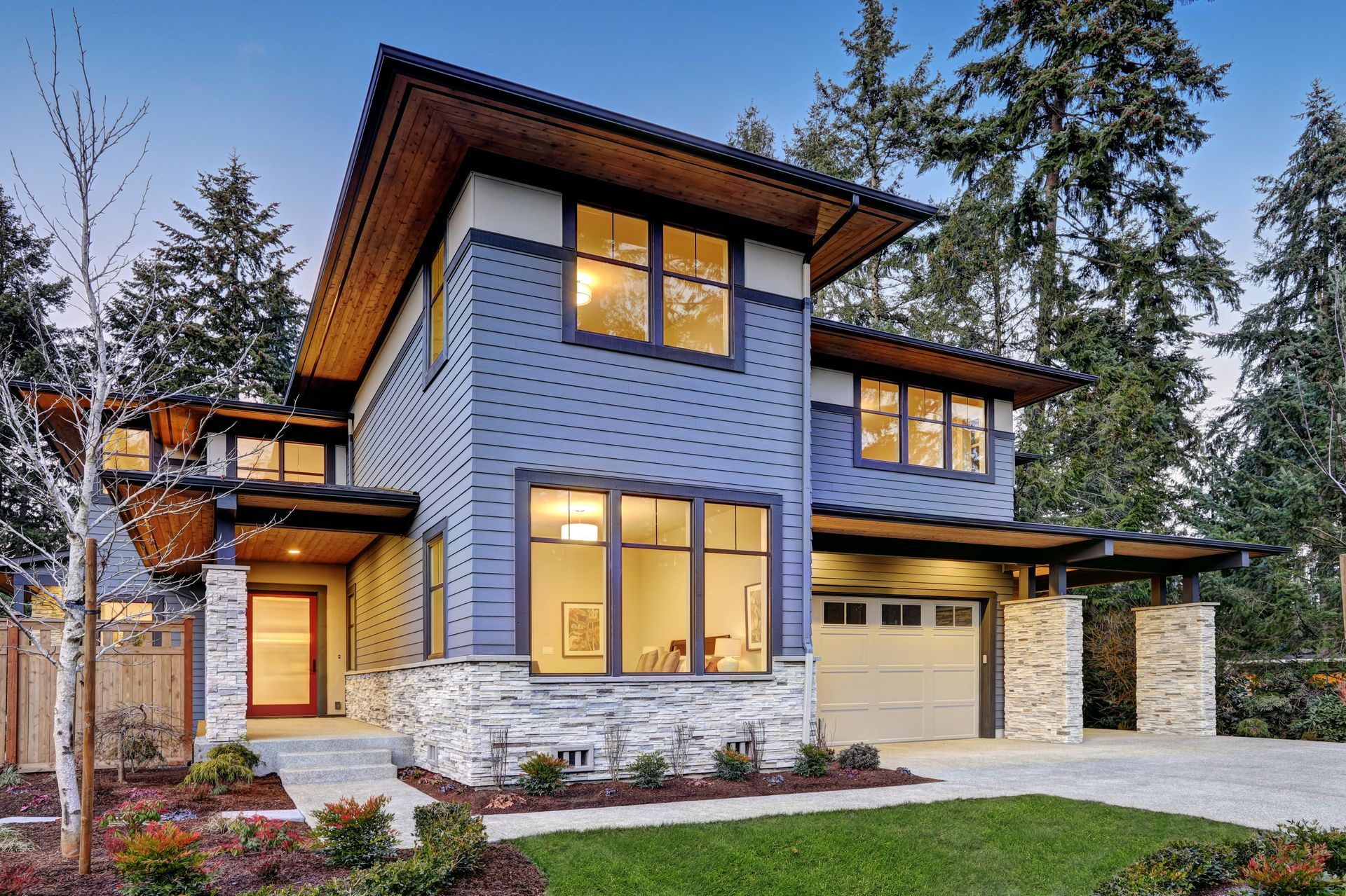 Two-story modern blue house with stone accents, large windows, and garage. Green lawn, trees.