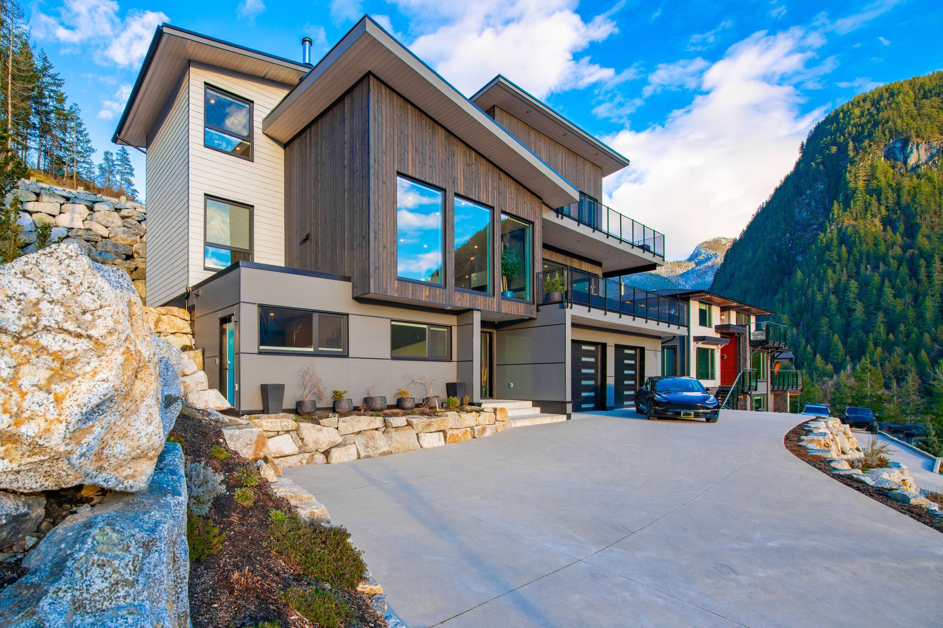 Modern house with gray and brown siding, large windows, and a mountain backdrop.