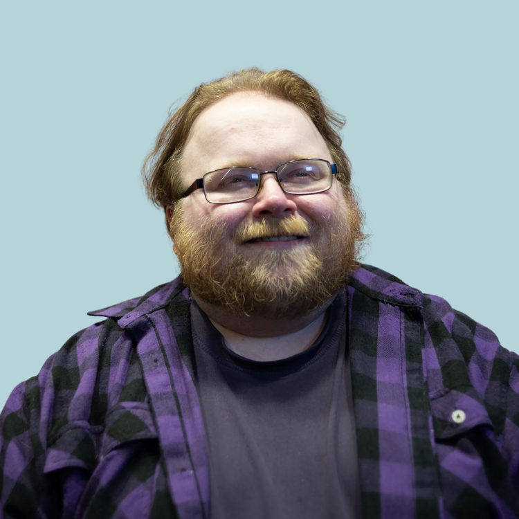Man with beard and glasses wearing purple plaid shirt, smiling.
