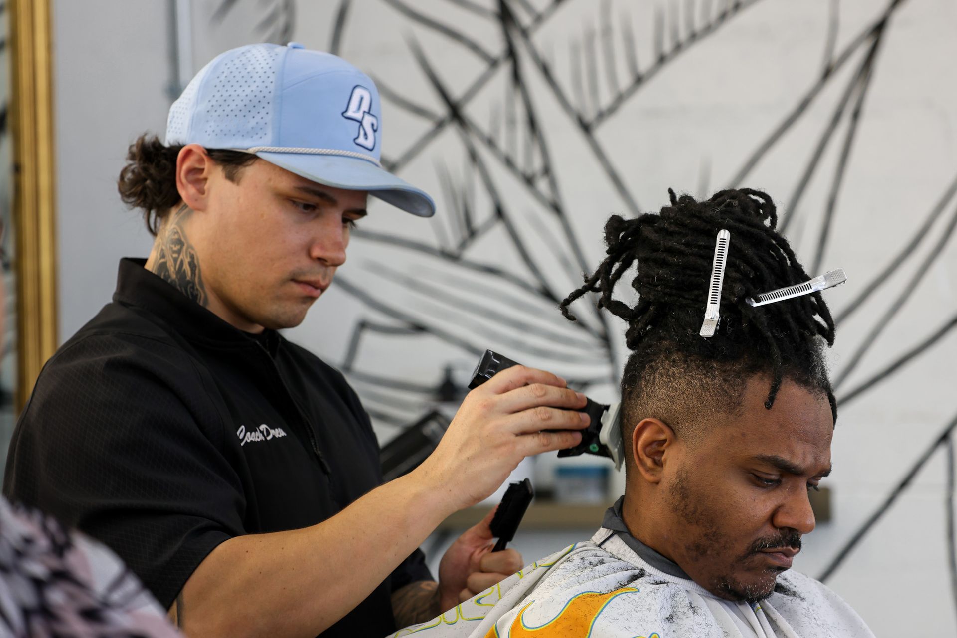 A barber wearing a light blue cap uses clippers to trim the hair of a client with dreadlocks in a barbershop.