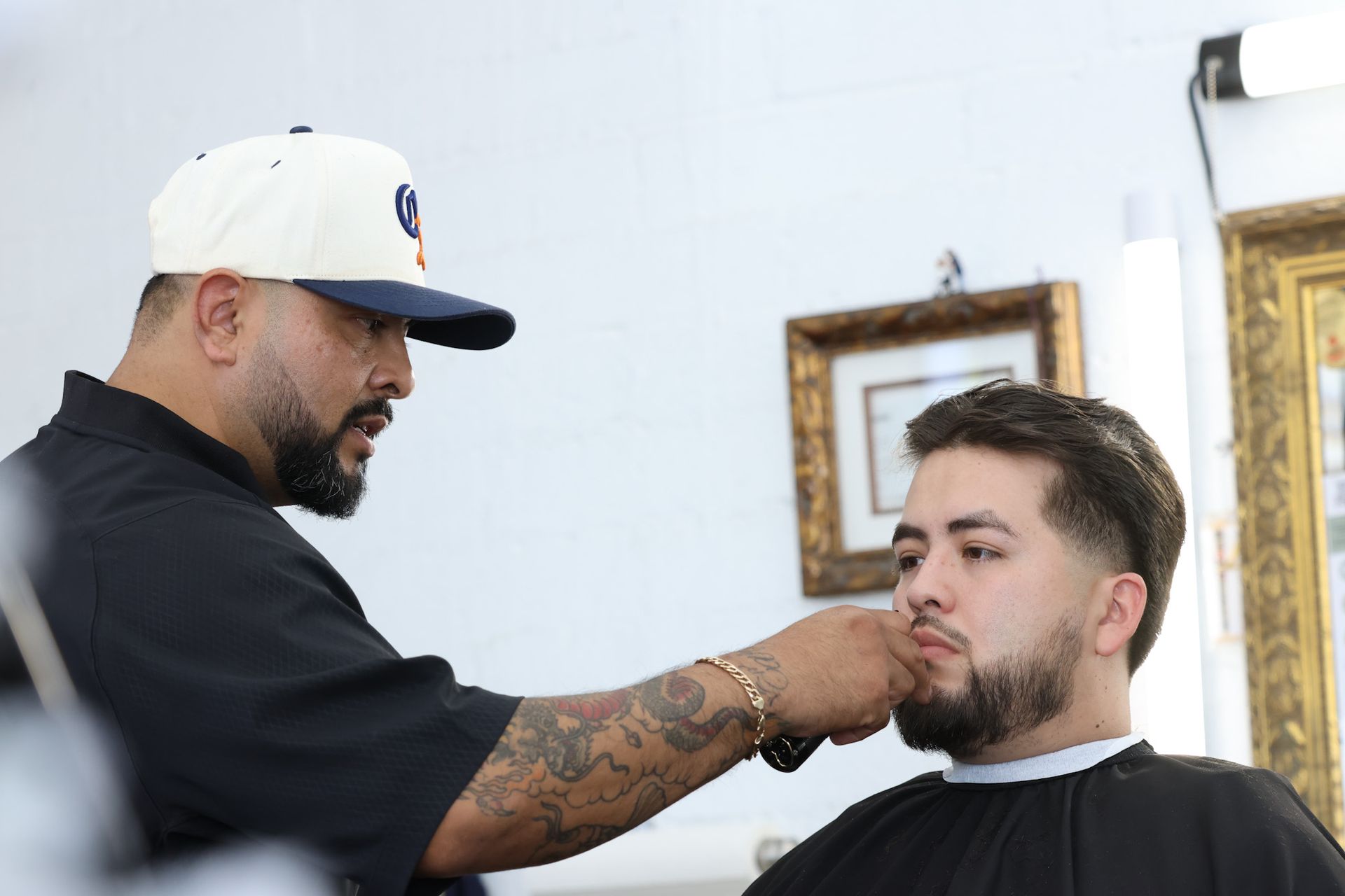 A barber with a tattooed arm uses an electric trimmer to shape a client's beard in a salon.