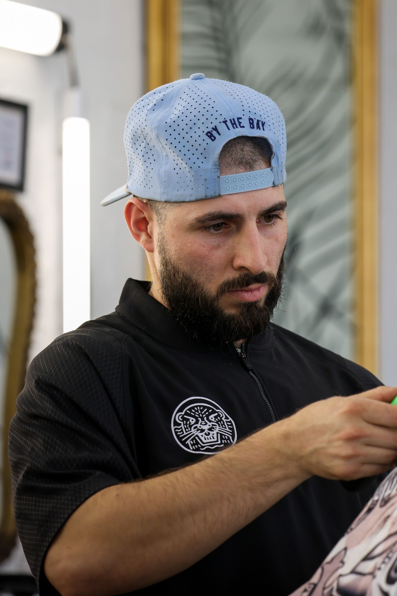 A person wearing a light blue cap and black barber's cape is focused on cutting a client's hair in a salon.