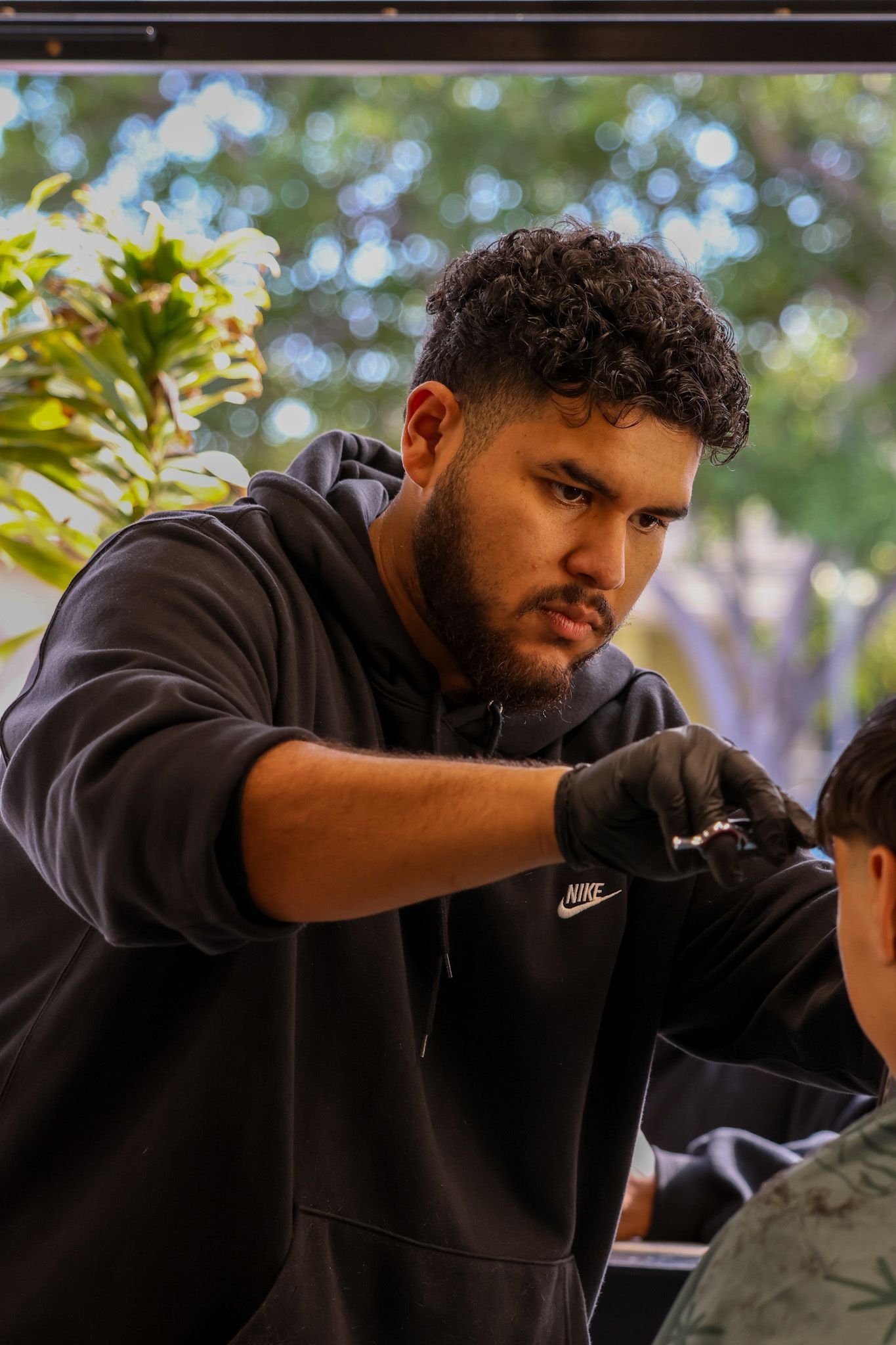 A barber in a black hoodie and black gloves carefully trims a client's hair with scissors.
