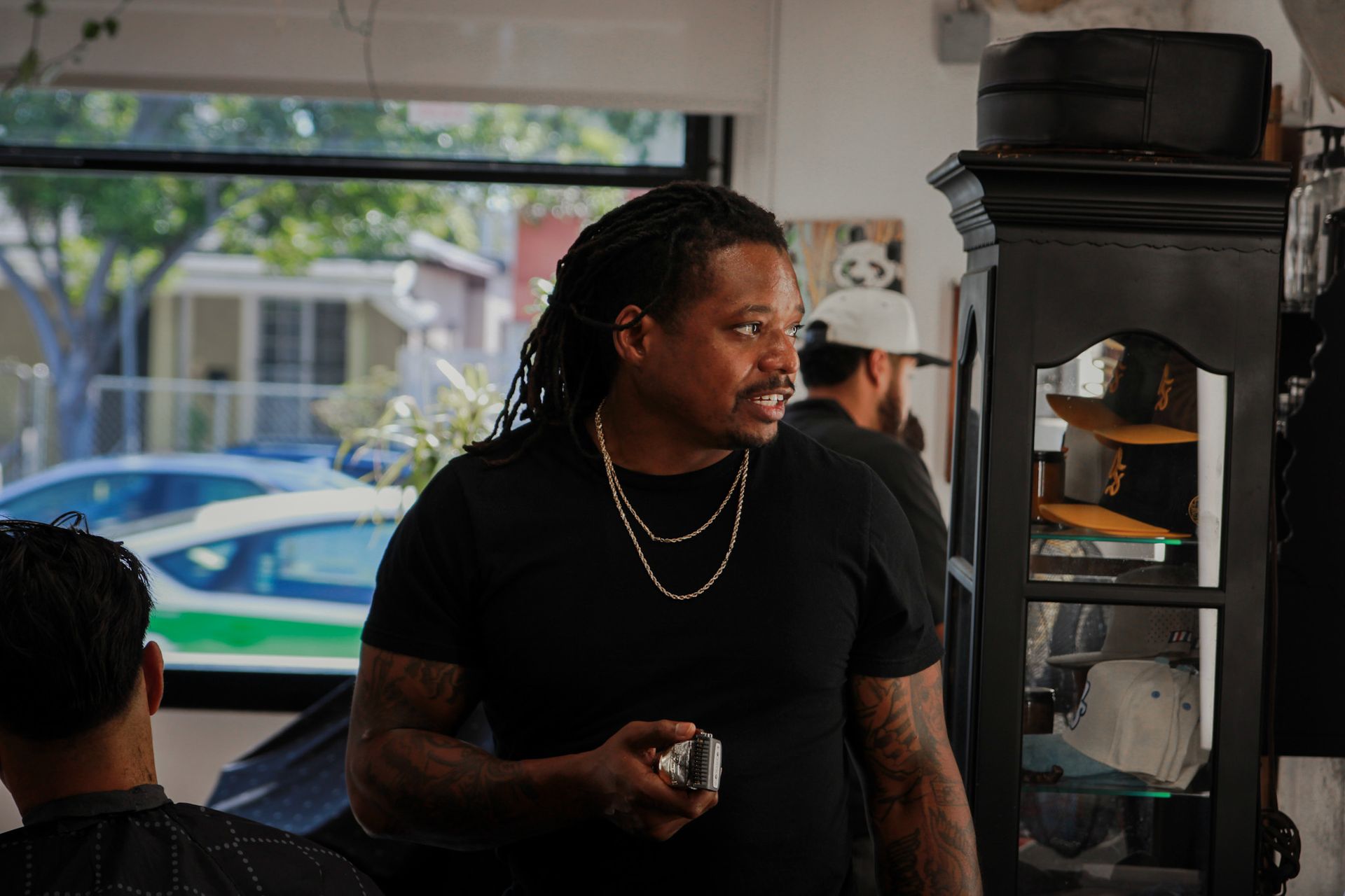 A barber with locs holding clippers in a shop, with a customer visible in the foreground and another person in the back.