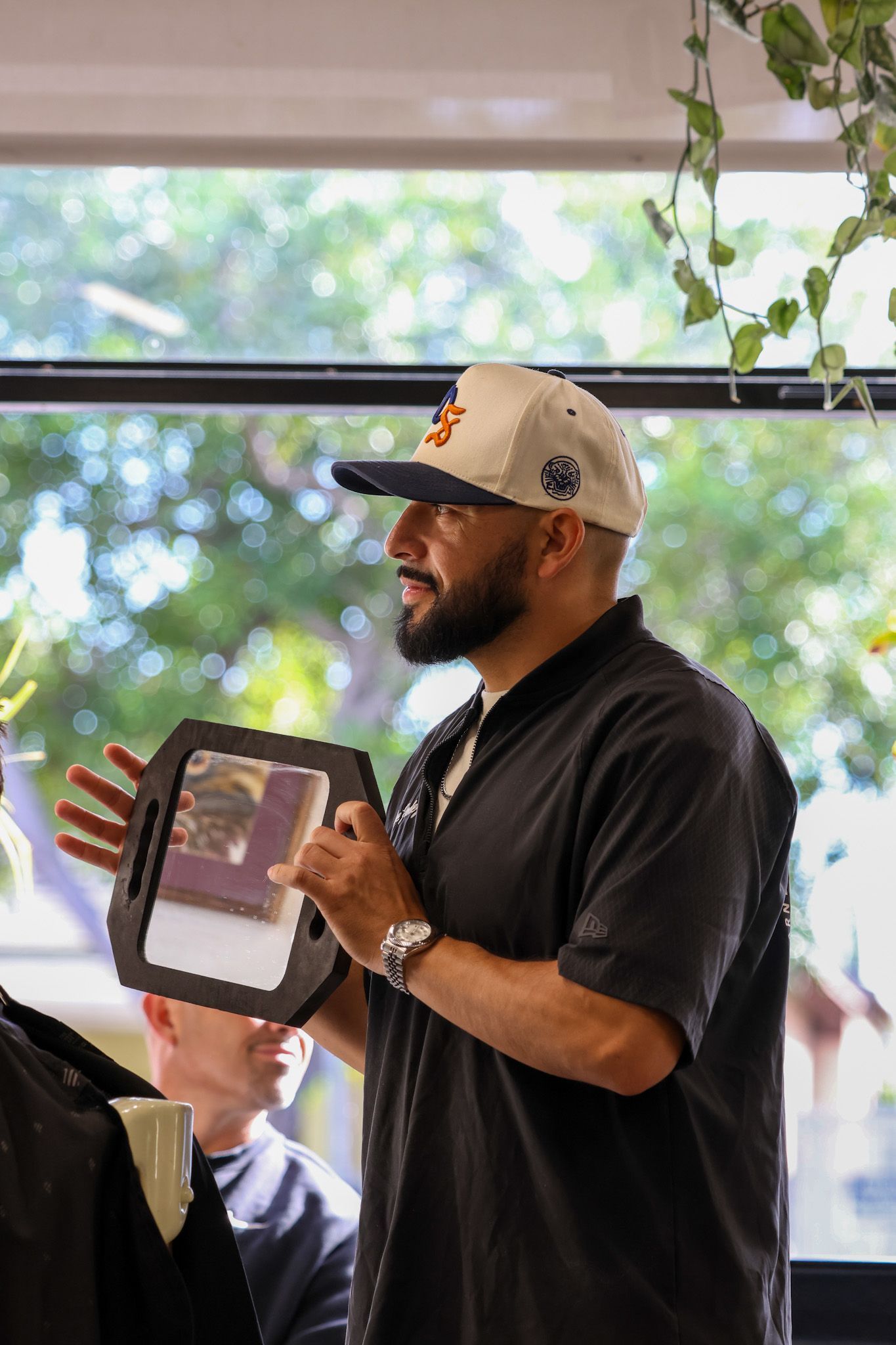 A barber in a cap and black shirt holds a handheld mirror for a client in a bright, plant-filled salon.