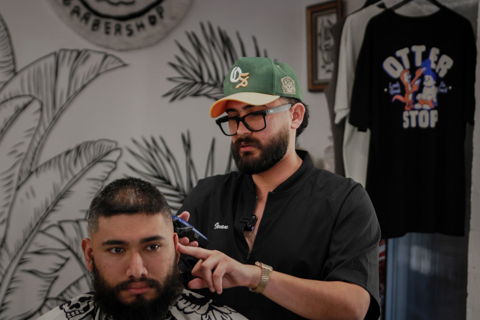 A barber in a cap and black shirt carefully trims a client’s hair in a salon with a tropical wall mural.
