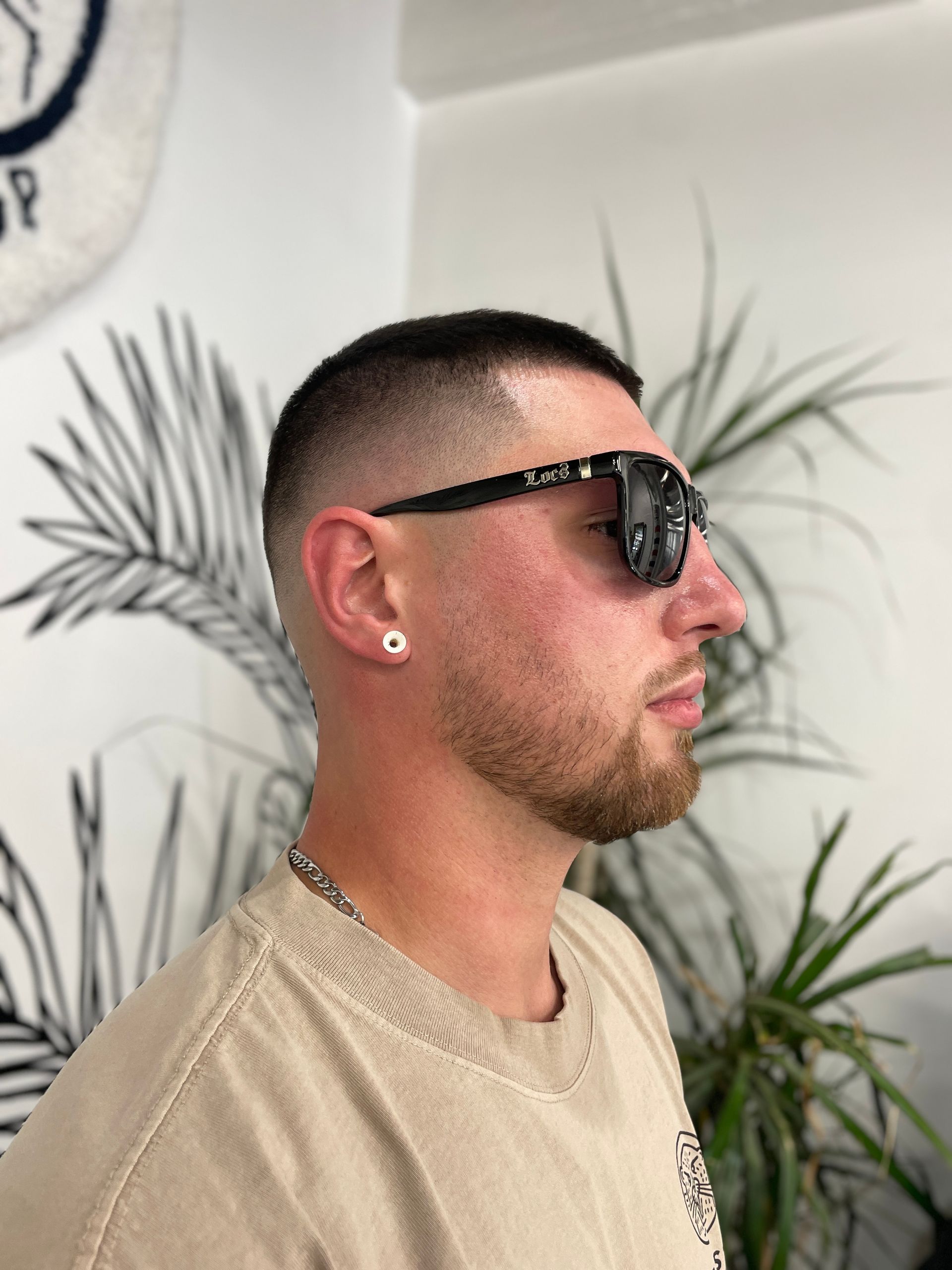 A side-profile shot of a person with a skin-fade haircut and beard, wearing dark sunglasses and a beige t-shirt.