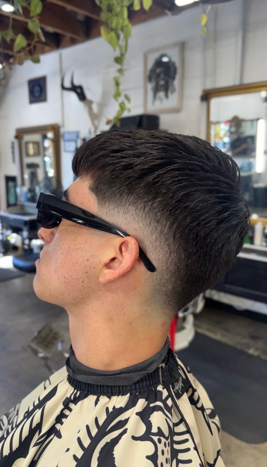 Side profile of a person in a barbershop wearing black glasses and showcasing a textured crop haircut with a skin fade.