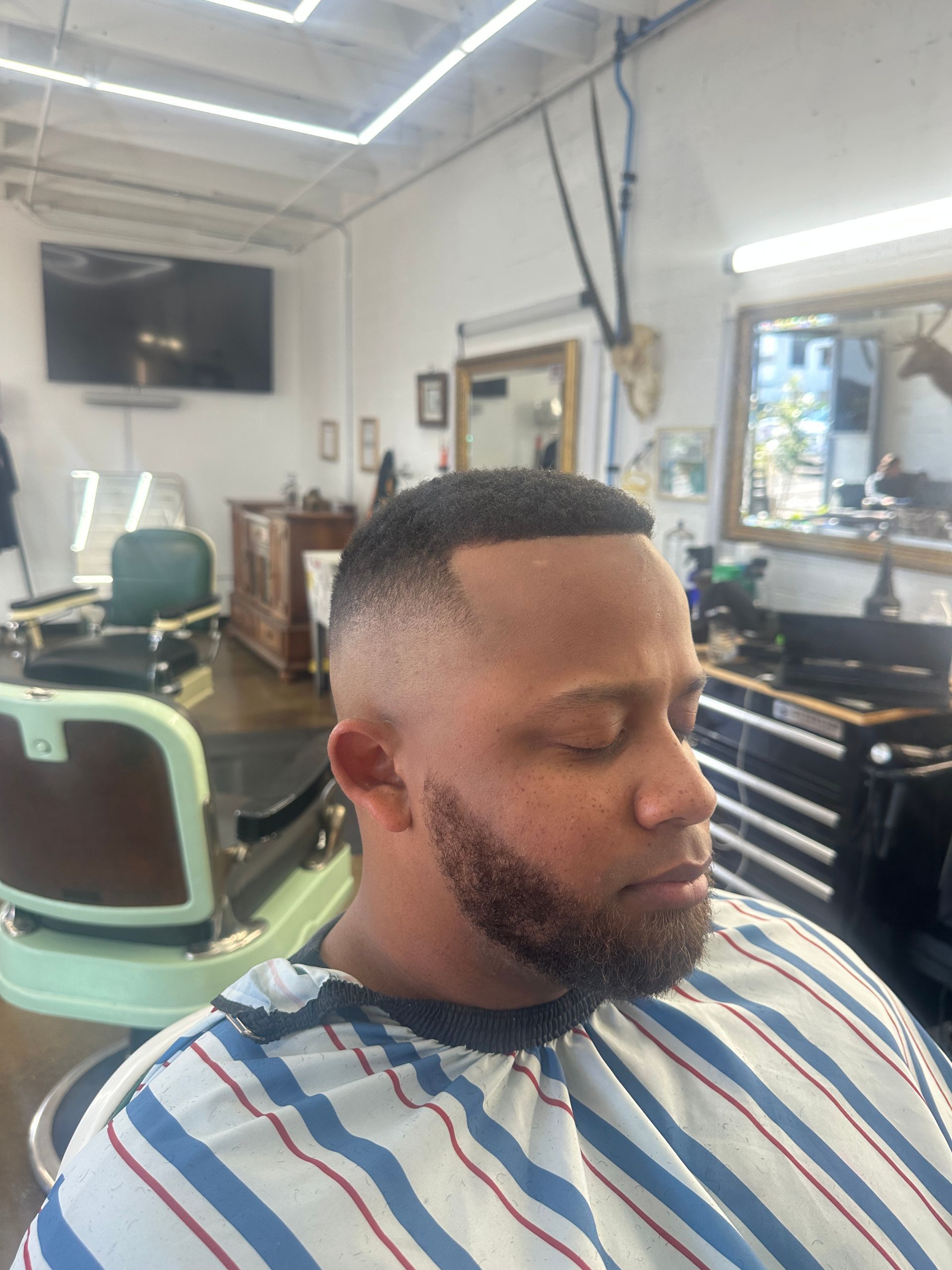 A person with a clean-cut fade hairstyle and trimmed beard, wearing a striped barber cape in a professional salon.
