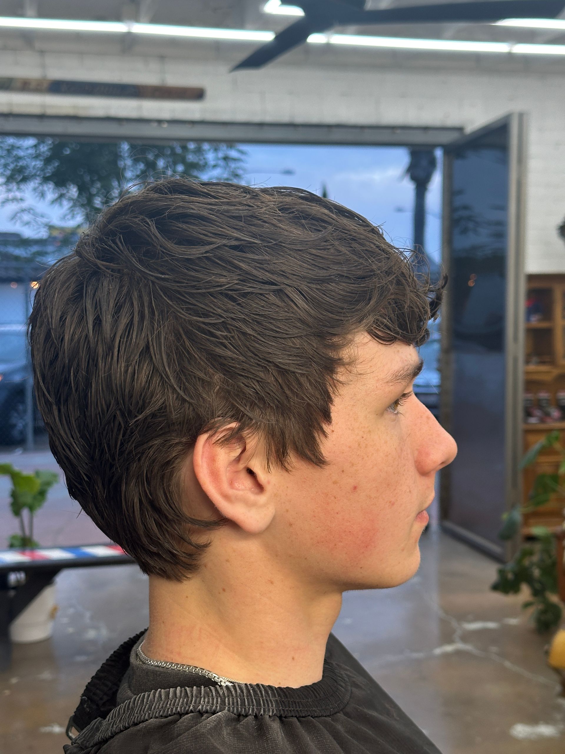 A side profile of a person with wavy, dark brown hair in a salon setting, wearing a black barber cape.