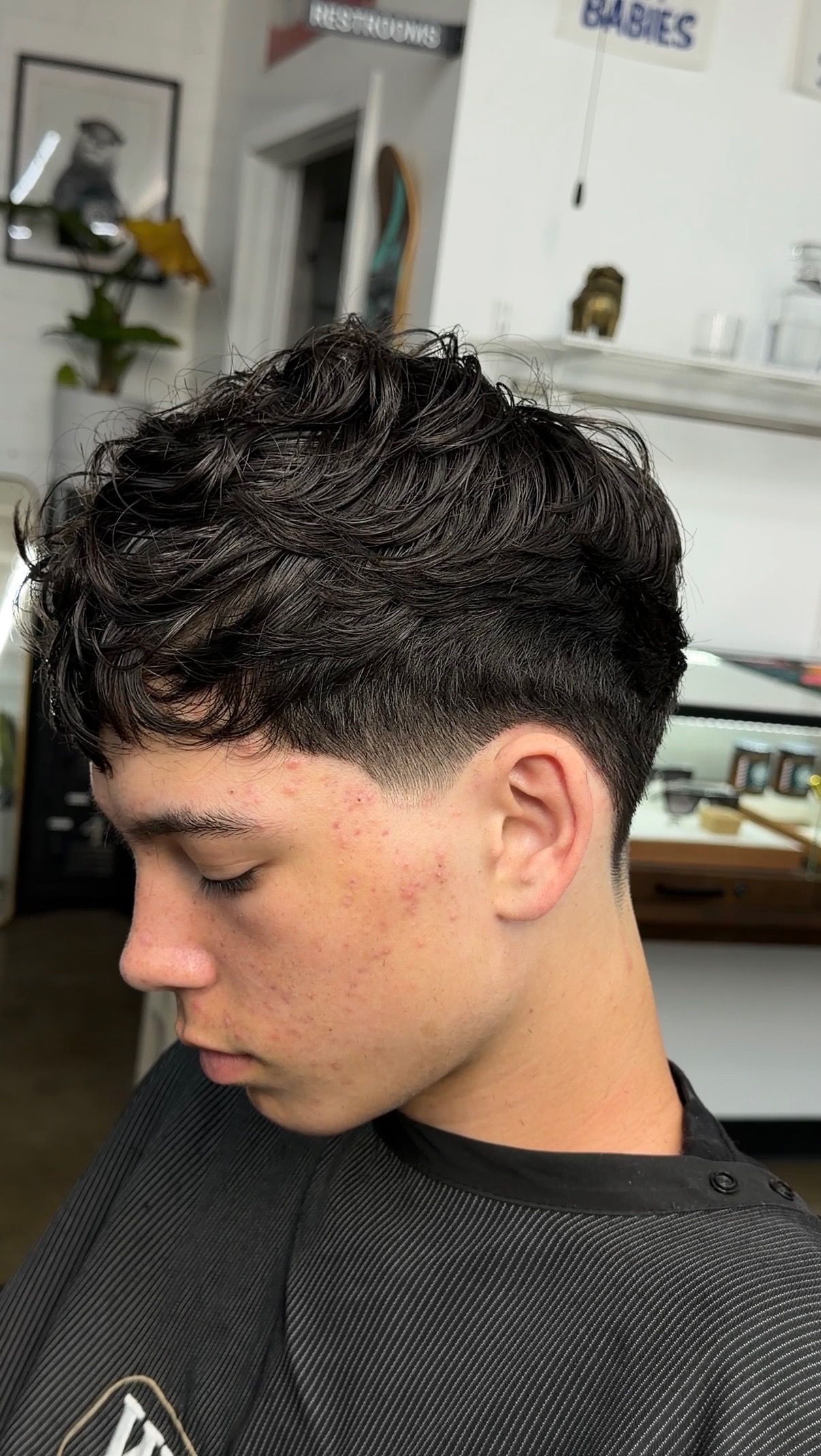 A person with short, curly dark hair and a faded haircut in a hair salon setting, shown in profile.