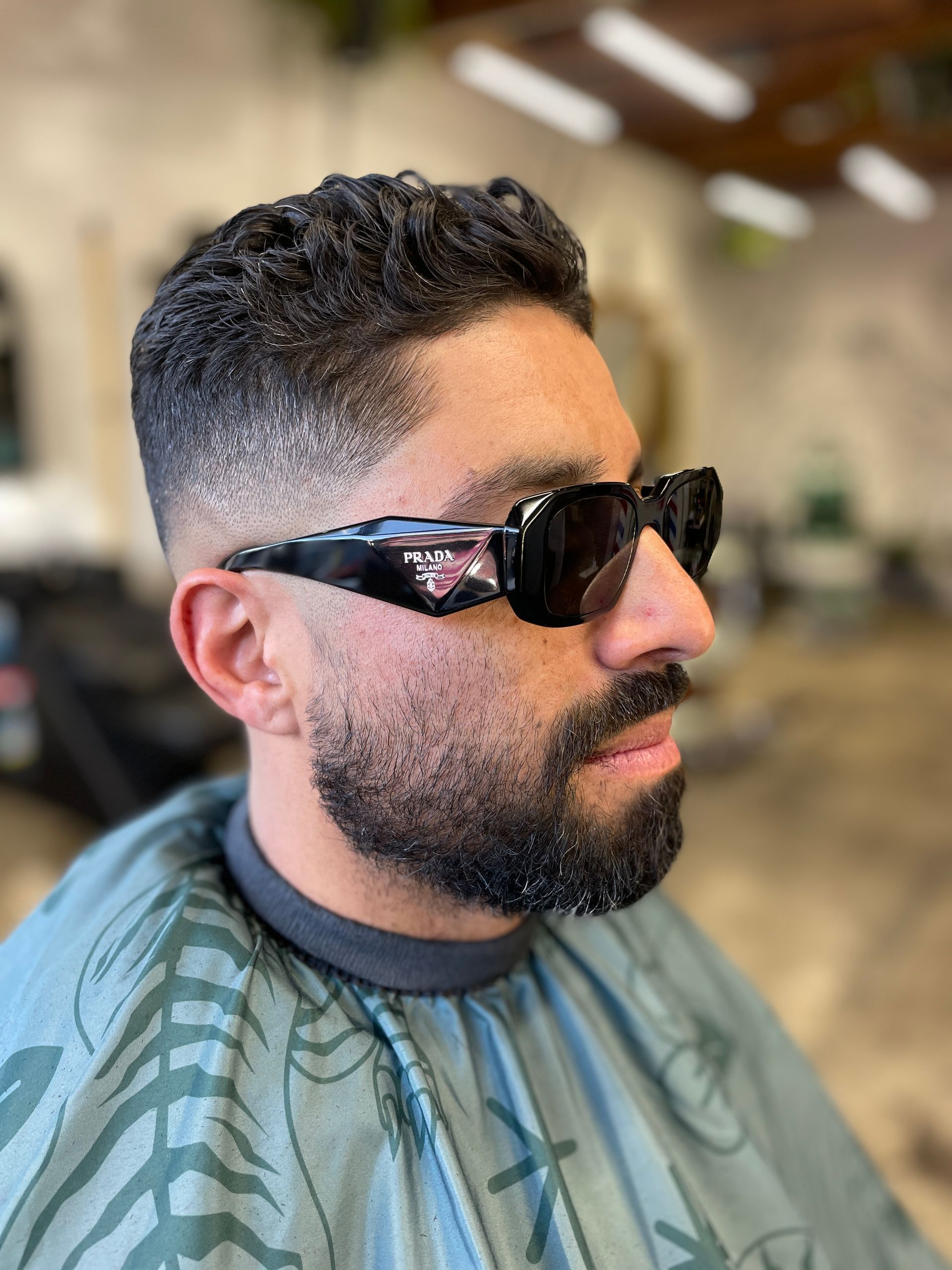 A person with a close fade haircut and a groomed beard wearing black sunglasses, seated in a hair salon.