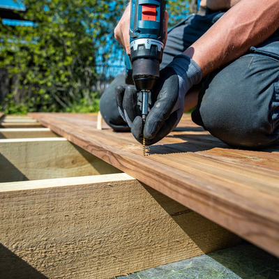 Person using a power drill to screw a board onto a wooden deck frame, outdoors, sunny day.