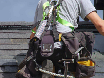 Roofer wearing a harness and tool belt on a shingled roof, preparing to work.