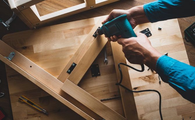 Hands using a blue power drill to assemble a wooden frame on a workbench.
